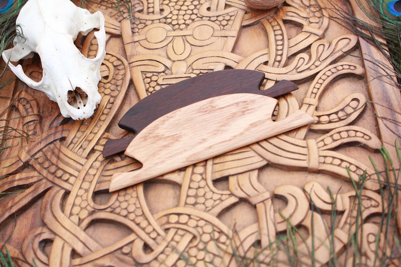 May include: Two wooden hair combs, one dark brown and one light brown, rest on a carved wooden surface with intricate patterns. A skull is visible in the upper left corner. The combs are designed with a unique shape, suggesting a Viking or Norse theme.