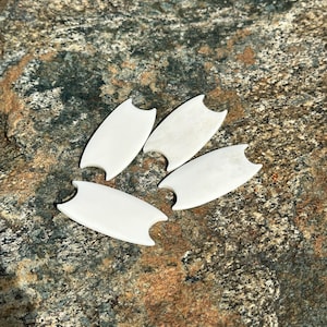 Op de afbeelding: Vier crèmekleurige, surfboard-vormige objecten met gebogen randen en een gekerfd ontwerp aan elk uiteinde. Ze zijn gerangschikt op een getextureerd, veelkleurig stenen oppervlak, wat een natuurlijke en aardse esthetiek creëert.