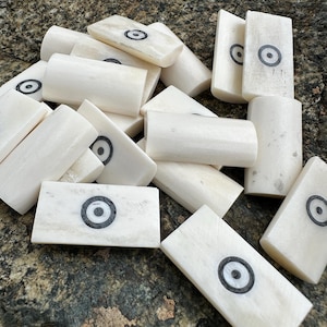May include: A collection of rectangular, off-white, smooth-surfaced pieces, each featuring a centred black and white circular bullseye or target pattern. The items are arranged in a scattered pile, displaying a consistent curved, cylindrical shape.