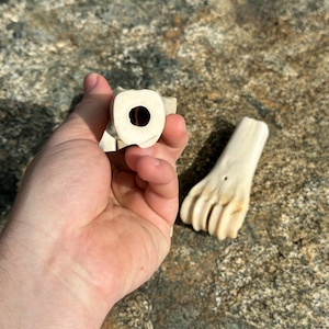 May include: Close-up of two carved bone objects. One object is held in a hand, showing a circular opening. The other object is a carved bone foot. The objects are off-white and appear to be handmade.