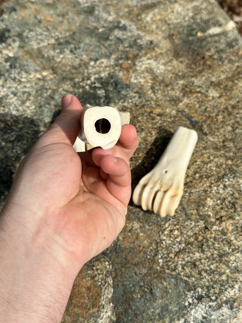 May include: Close-up of two carved bone objects. One object is held in a hand, showing a circular opening. The other object is a carved bone foot. The objects are off-white and appear to be handmade.