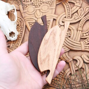 May include: Two wooden leaf-shaped hair combs, one dark brown and one light brown, are held in a hand. A skull and carved wooden background are visible. Peacock feathers and grass-like plants are in the background.