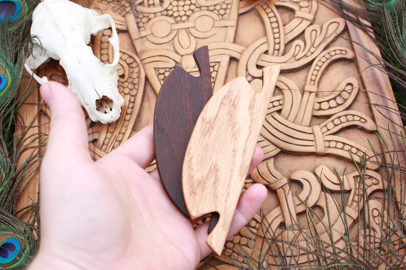 May include: Two wooden leaf-shaped hair combs, one dark brown and one light brown, are held in a hand. A skull and carved wooden background are visible. Peacock feathers and grass-like plants are in the background.