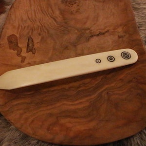 May include: A white bone tool with a pointed end and three black circles on the handle. The tool is resting on a wooden surface.
