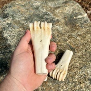 May include: Two off-white, carved bone objects, possibly decorative or functional, are held against a natural stone background. Each piece has a handle-like base and a splayed, finger-like top with a small hole.