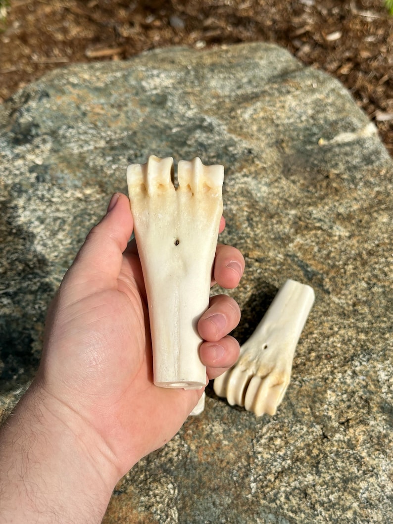 May include: Two off-white, carved bone objects, possibly decorative or functional, are held against a natural stone background. Each piece has a handle-like base and a splayed, finger-like top with a small hole.