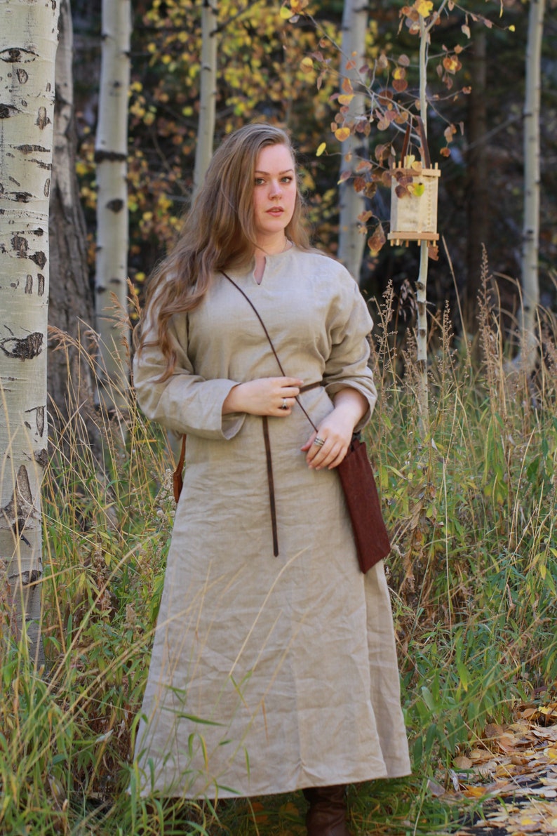 Puede incluir: Una mujer con un vestido largo de lino marr&oacute;n claro con un cintur&oacute;n de cuero marr&oacute;n y un bolso de cuero marr&oacute;n. Est&aacute; de pie en un bosque con &aacute;rboles altos y hierba verde.