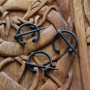 May include: Three antique-looking metal brooches with a circular design and a pin. The brooches are arranged on a wooden surface with intricate carvings.