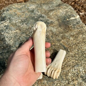 May include: Two pieces of carved bone, one held in a hand. The bone pieces are off-white and appear to be carved into a cylindrical shape and a foot shape. The background is a large, gray rock.