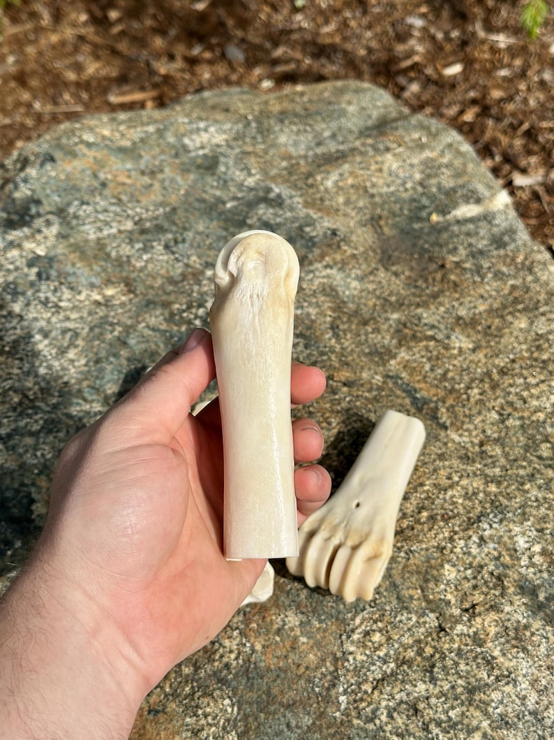 May include: Two pieces of carved bone, one held in a hand. The bone pieces are off-white and appear to be carved into a cylindrical shape and a foot shape. The background is a large, gray rock.