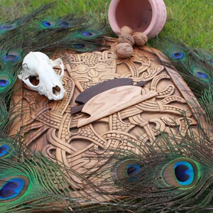 May include: A wooden carving with intricate designs, a skull, and two wooden objects are displayed. Peacock feathers with blue and green accents surround the carving. A small pink pot and walnuts are also present, all set on a bed of green grass.