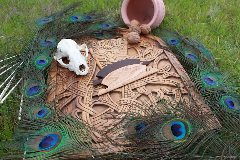 May include: A wooden carving with intricate designs, a skull, and two wooden objects are displayed. Peacock feathers with blue and green accents surround the carving. A small pink pot and walnuts are also present, all set on a bed of green grass.