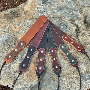 May include: A collection of six leather camera straps in various colors, including brown, black, and green. Each strap features a decorative design with silver rivets and a black cord for attachment. The straps are arranged on a rock surface.