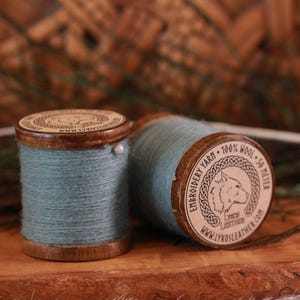 May include: Two wooden spools wrapped with light blue 100% wool embroidery yarn. Each spool features a circular label with a wolf illustration, the text EMBROIDERY YARN, 100% WOOL, 50 METER, and the website address WWW.LYKOSLEATHER.COM.