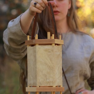 Puede incluir: Una mujer sostiene una linterna de madera con una correa de cuero. La linterna est&aacute; hecha de madera y tiene un material parecido al pergamino para los lados. La linterna est&aacute; colgada de la correa.