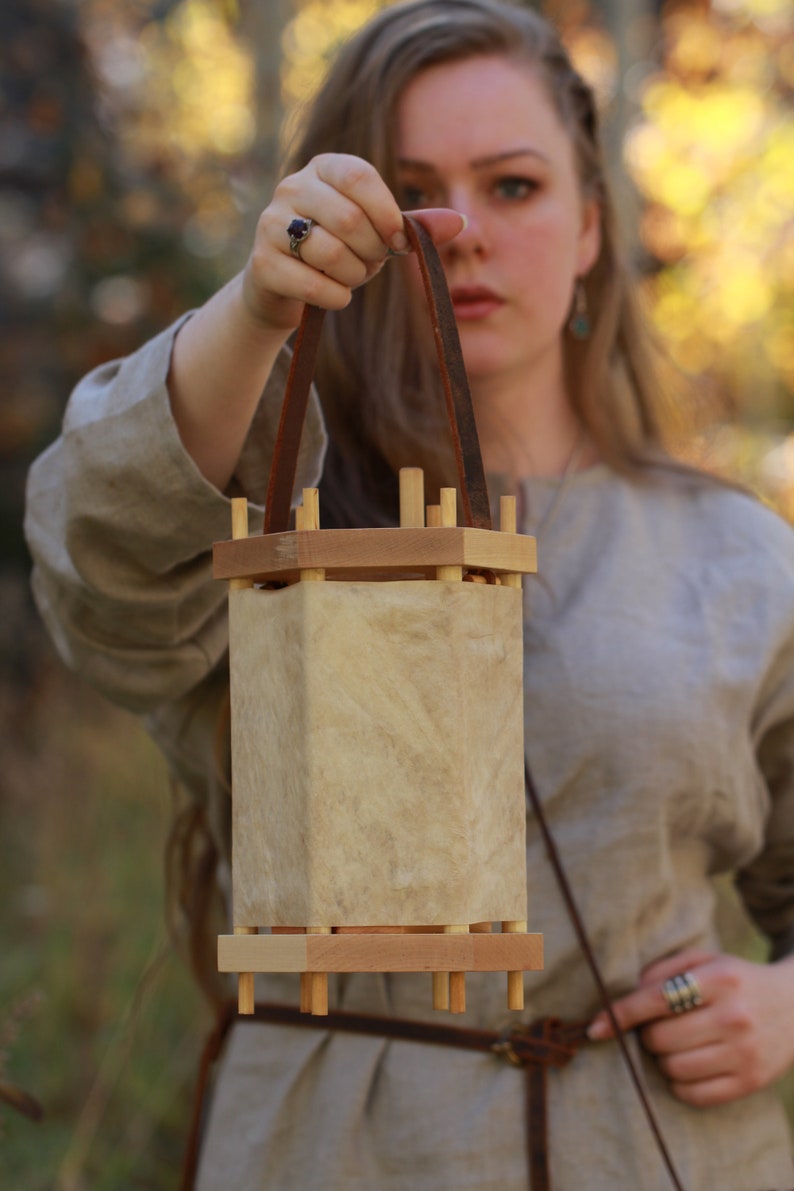 Puede incluir: Una mujer sostiene una linterna de madera con una correa de cuero. La linterna est&aacute; hecha de madera y tiene un material parecido al pergamino para los lados. La linterna est&aacute; colgada de la correa.