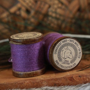 Puede incluir: Dos carretes de hilo de bordar morado, uno con un carrete de madera y el otro con una etiqueta que dice "Hilo de bordar - 100% lana - 50 metros - www.lykosleather.com".
