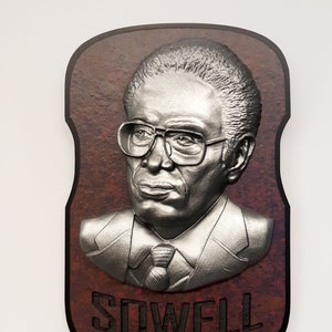 May include: A silver relief sculpture of a man in glasses and a suit, mounted on a dark brown plaque. The plaque has a reddish-brown textured background and the name "SOWELL" is carved at the bottom.