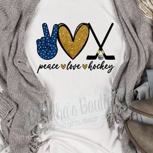 May include: White t-shirt with a peace sign, heart, and hockey sticks. The peace sign is blue with white polka dots. The heart is gold glitter. The hockey sticks are black with a gold puck. The text "peace love hockey" is written below the design.
