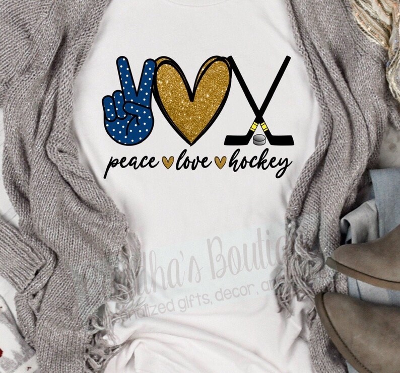May include: White t-shirt with a peace sign, heart, and hockey sticks. The peace sign is blue with white polka dots. The heart is gold glitter. The hockey sticks are black with a gold puck. The text "peace love hockey" is written below the design.