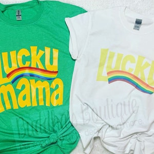 May include: Two t-shirts with the text "Lucky Mama" in yellow with a rainbow arc above the text. The t-shirt on the left is green and the t-shirt on the right is white.