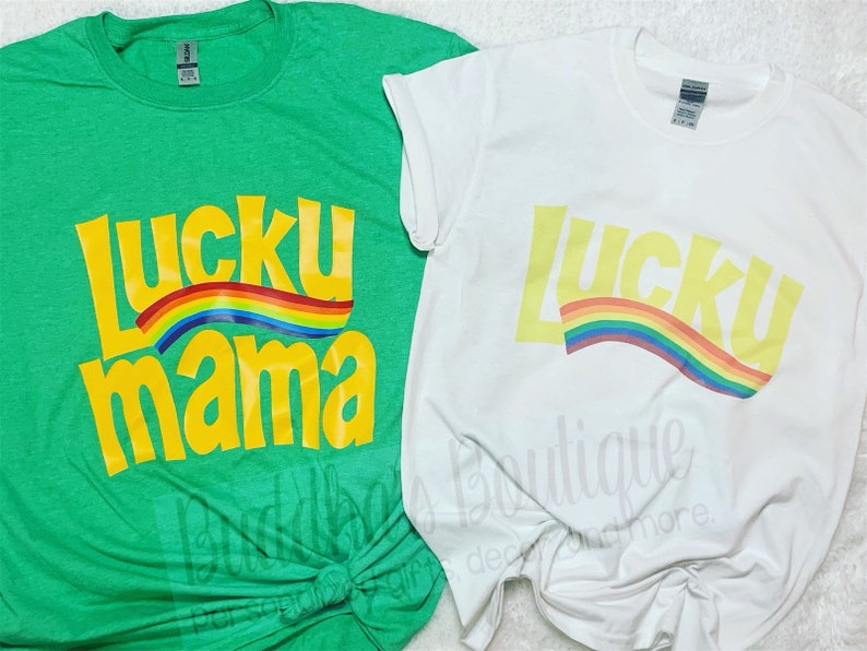 May include: Two t-shirts with the text "Lucky Mama" in yellow with a rainbow arc above the text. The t-shirt on the left is green and the t-shirt on the right is white.