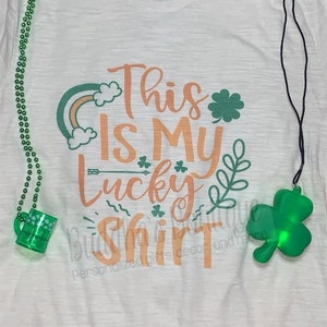 May include: A white t-shirt with the text "This is my lucky shirt" in orange and green. The shirt has a rainbow, shamrocks, and leaves printed on it.