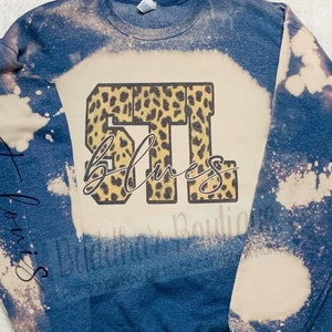 May include: A blue sweatshirt with a bleach-splattered design. The sweatshirt has a leopard print graphic with the text "STL Blues" on the front.