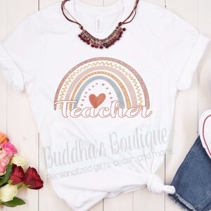 May include: White t-shirt featuring a rainbow design and the word "Teacher" in brown lettering. The rainbow is made up of different shades of brown, beige, and blue. A small red heart is in the centre of the rainbow. The text "Buddha's Boutique" is at the bottom of the shirt.