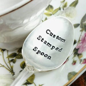 May include: A silver spoon with the words "Custom Stamped Spoon" engraved on the front. The spoon is resting on a white saucer with a floral pattern.