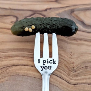May include: A silver fork with the words "i pick you" engraved on the handle. A dill pickle is balanced on the tines of the fork.
