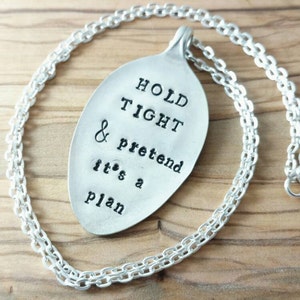May include: A silver-colored spoon pendant necklace with the words "HOLD TIGHT & pretend it's a plan" stamped on the spoon. The necklace has a chain with a lobster clasp.
