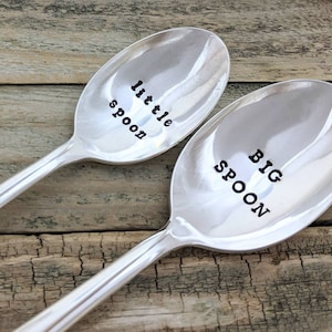 May include: Two silver spoons with the words "little spoon" and "big spoon" engraved on them. The spoons are lying on a wooden surface.