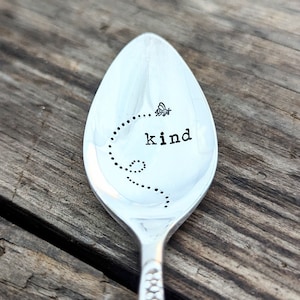 May include: A silver spoon with the word "kind" engraved on it. A small bee design is above the word. The spoon is on a wooden surface.