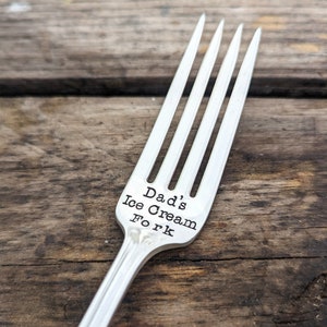 May include: A silver fork with the words "Dad's Ice Cream Fork" engraved on the handle. The fork is lying on a wooden surface.