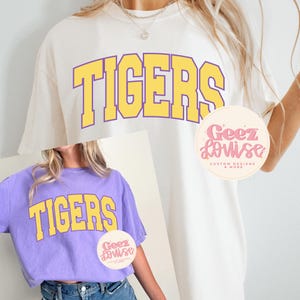 Puede incluir: Camisetas crema y moradas con la palabra "TIGERS" en letras amarillas. Las camisetas tienen un estilo vintage y universitario. Un logotipo circular con el texto "Geez Louise Custom Designs & More" es visible.