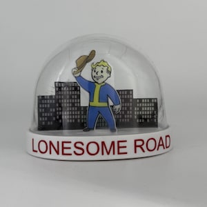 May include: A clear snow globe with a white base featuring the text "Lonesome Road." Inside, a cartoon figure in a blue jumpsuit and a brown hat stands before a cityscape. The figure is waving.
