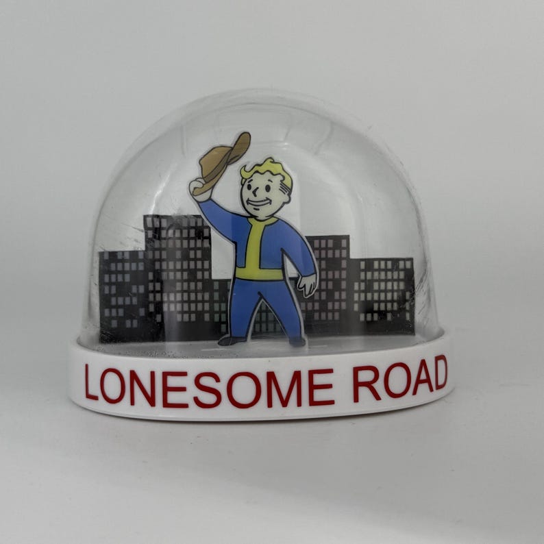 May include: A clear snow globe with a white base featuring the text "Lonesome Road." Inside, a cartoon figure in a blue jumpsuit and a brown hat stands before a cityscape. The figure is waving.