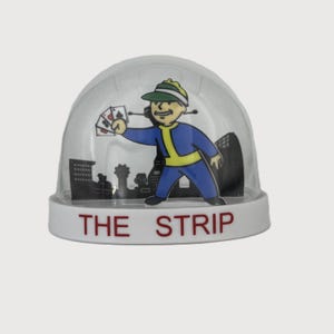 May include: A clear snow globe with a white base featuring a cartoon character in a blue jumpsuit and green cap holding playing cards. The base reads "THE STRIP" in red letters, with a city skyline backdrop.