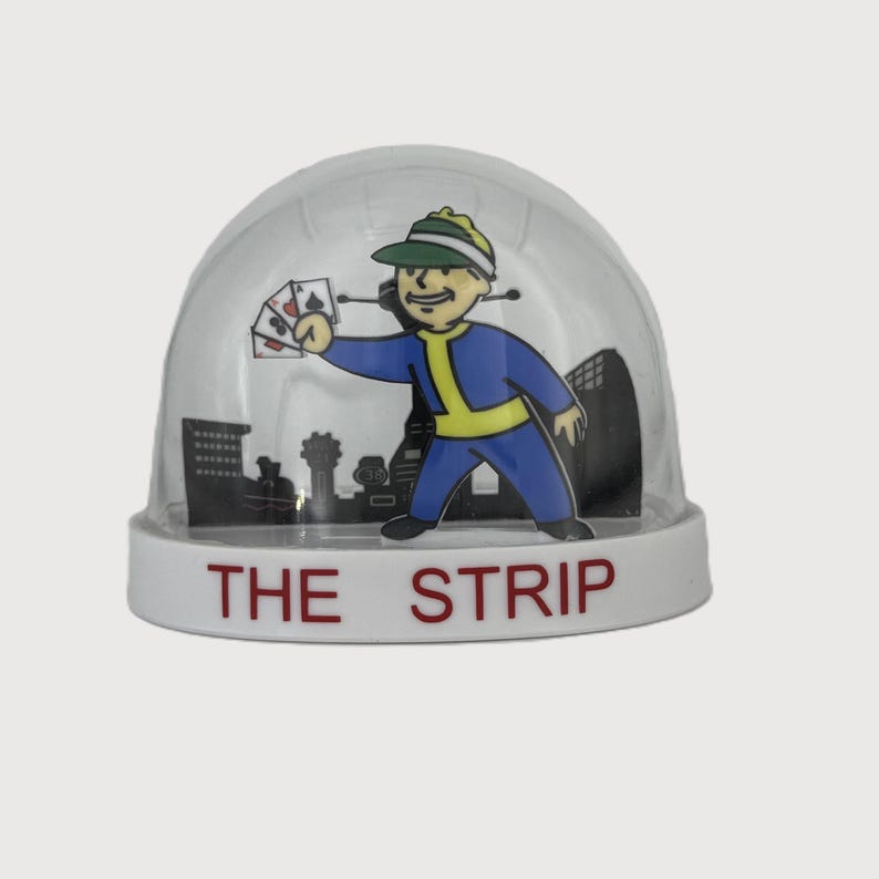 May include: A clear snow globe with a white base featuring a cartoon character in a blue jumpsuit and green cap holding playing cards. The base reads "THE STRIP" in red letters, with a city skyline backdrop.