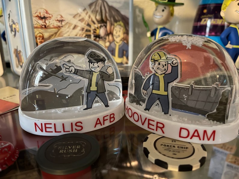 May include: Two snow globes with white bases and clear domes. One features a cartoon figure with the text "NELLIS AFB" in red. The other has a cartoon figure with the text "HOOVER DAM" in red. Both globes have a Fallout theme.
