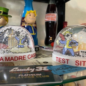 May include: Two snow globes with white bases and clear domes. One says "SIERRA MADRE CASINO" and the other says "TEST SITE". Each globe features a cartoon character in a blue and yellow jumpsuit. Other figurines and a Coca-Cola bottle are in the background.