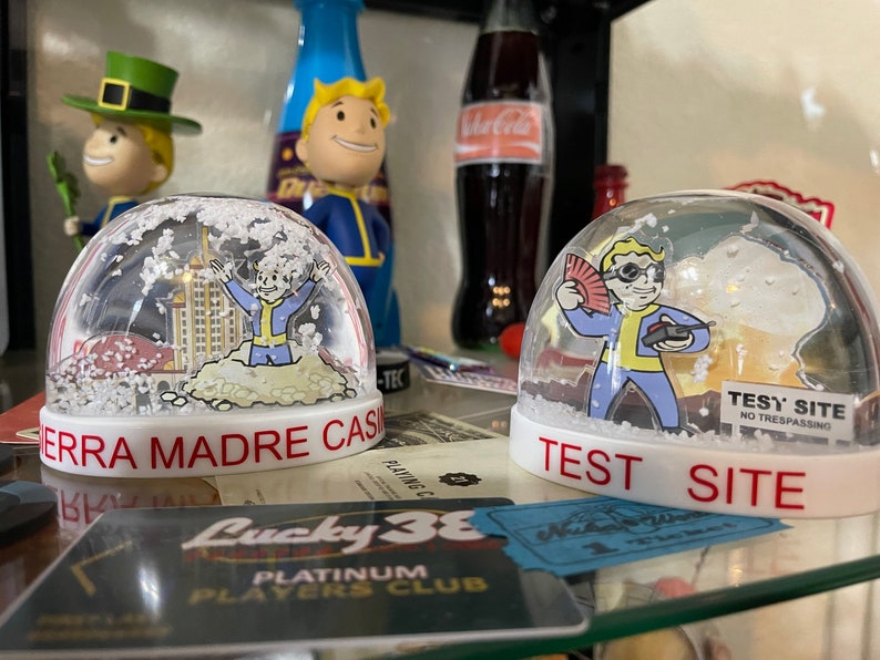 May include: Two snow globes with white bases and clear domes. One says "SIERRA MADRE CASINO" and the other says "TEST SITE". Each globe features a cartoon character in a blue and yellow jumpsuit. Other figurines and a Coca-Cola bottle are in the background.
