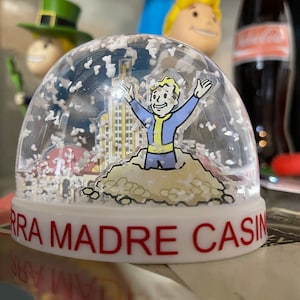 May include: A clear snow globe featuring a cartoon character with arms raised, wearing a blue and yellow jumpsuit. The globe has a building and snow inside. The base is white with the words "SIERRA MADRE CASINO" in red.