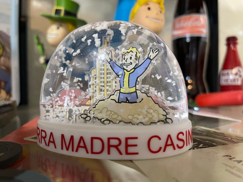 May include: A clear snow globe featuring a cartoon character with arms raised, wearing a blue and yellow jumpsuit. The globe has a building and snow inside. The base is white with the words "SIERRA MADRE CASINO" in red.