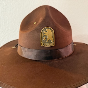 May include: Brown felt Ranger hat with a wide brim and a dark brown ribbon band. A gold-colored metal badge with a bear and star design is centered on the front, with the word "RANGER" below. The hat has a classic, outdoorsy style.