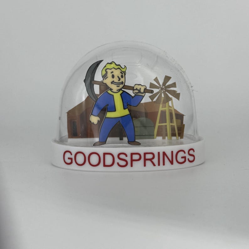 May include: A clear snow globe with a white base featuring the text "GOODSPRINGS" in red. Inside, a cartoon figure in blue and yellow holds a pickaxe, with a windmill and building in the background.