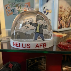 May include: A clear snow globe with a cartoon figure in a military uniform pointing towards a silhouette of an airplane. The base of the globe is white with the red text "NELLIS AFB". The background includes other decorative items.