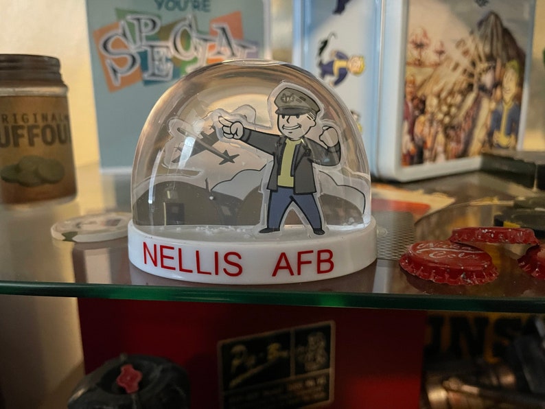 May include: A clear snow globe with a cartoon figure in a military uniform pointing towards a silhouette of an airplane. The base of the globe is white with the red text "NELLIS AFB". The background includes other decorative items.