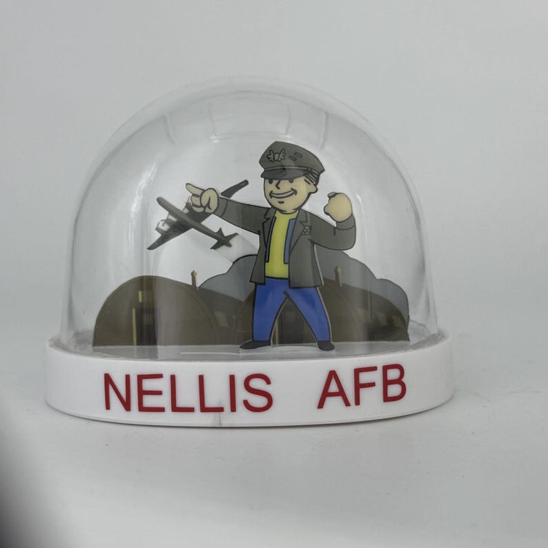 May include: A clear snow globe with a cartoon illustration of a man in a military uniform pointing at a plane. The base of the globe is white with the text "NELLIS AFB" in red.